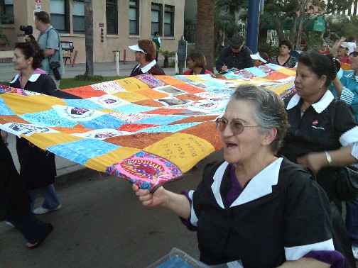Women of many ages and races walk together along a street, dressed in hotel uniforms, each holding part of the edge of a large quilt