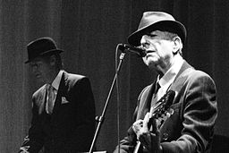 Photograph of an elderly pale-skinned man, wearing a suit and hat, singing into a microphone while playing a guitar.