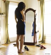A dark-haired woman in a black dress looking at herself in a full-length mirror