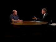 Charlie Rose sits across a table from Louis CK against a black background, gesturing during an interview